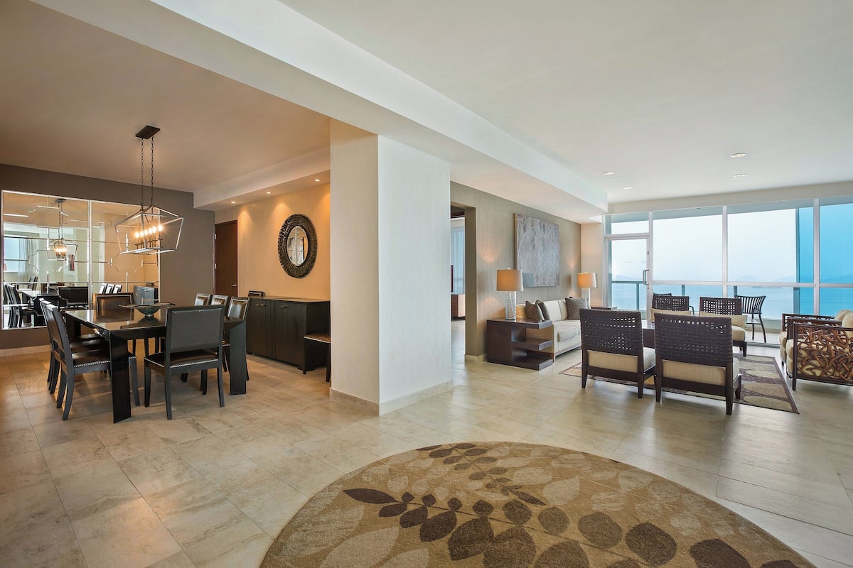 A spacious living area with a dining table, chairs, a sofa, and a large window offering a view.