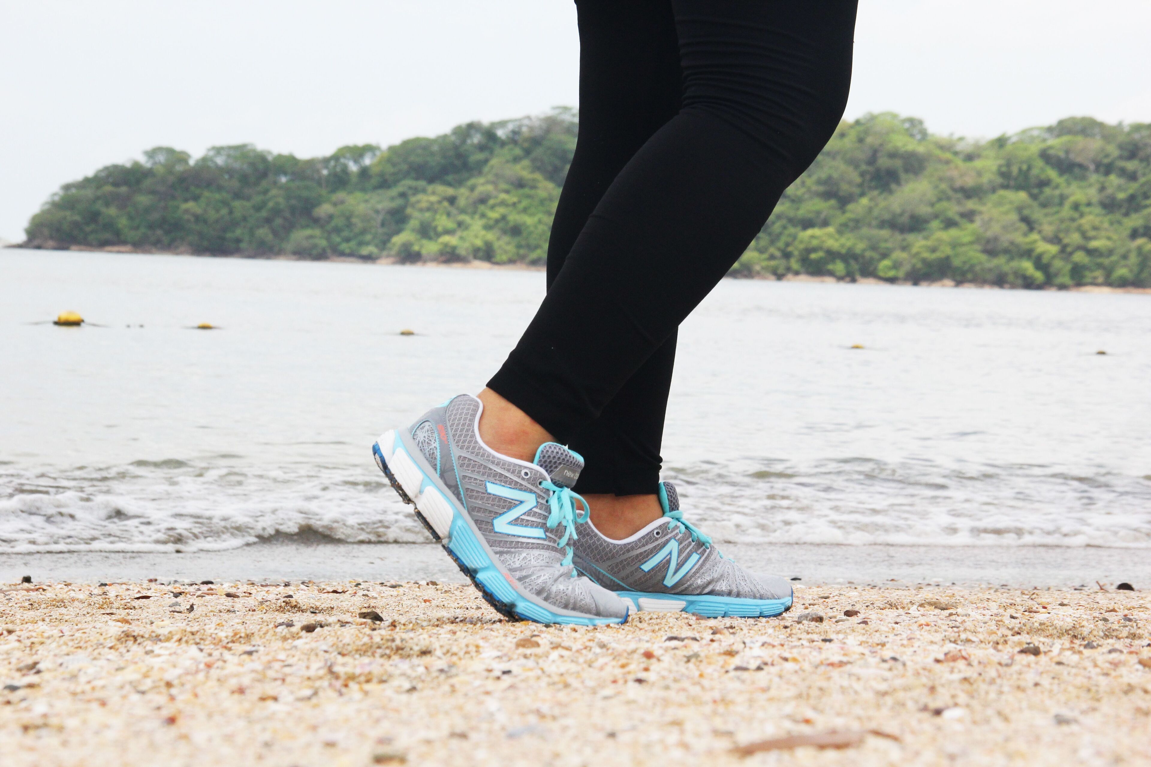 A close-up of a person's feet wearing grey and blue running shoes with the brand 'N' visible, standing on a sandy beach with water and greenery in the background.