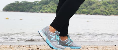 A close-up of a person's feet wearing grey and blue running shoes with the brand 'N' visible, standing on a sandy beach with water and greenery in the background.