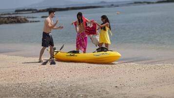 Escena de playa con un kayak amarillo, gente preparándose para actividades acuáticas y un mar tranquilo al fondo.