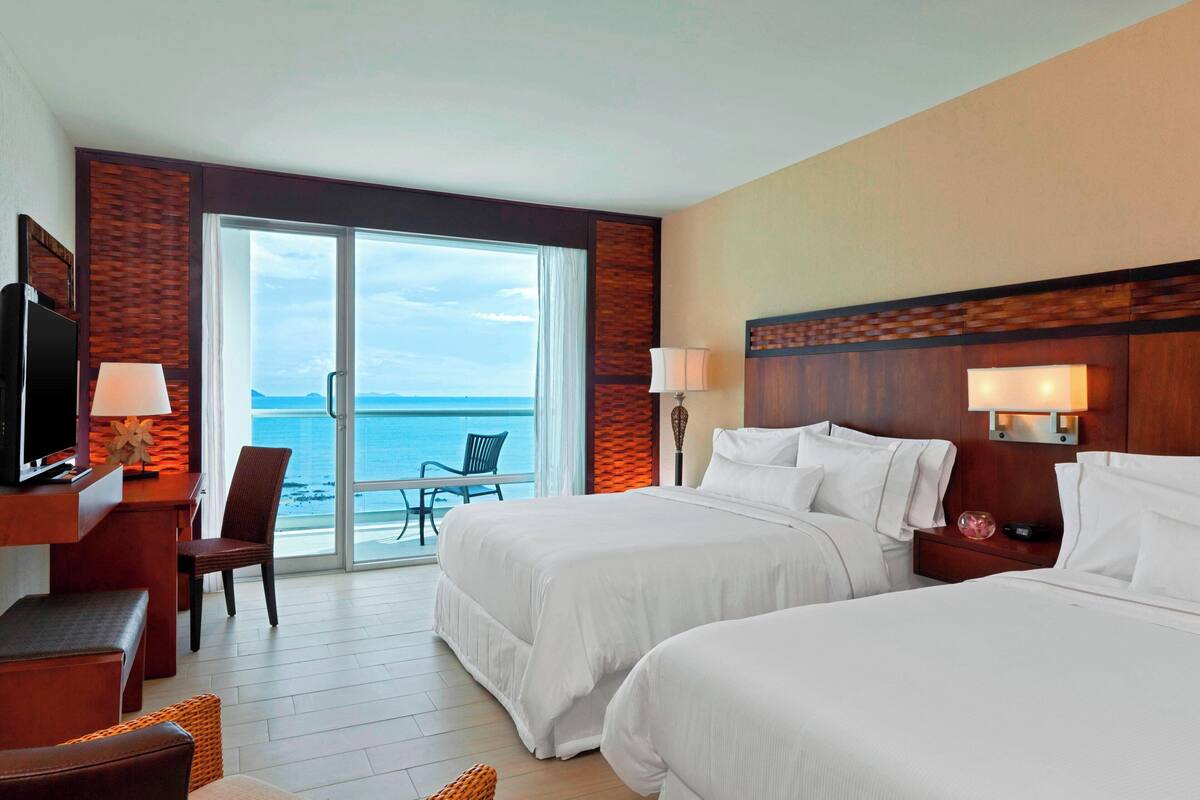 A hotel room with two beds, a desk with a TV, a chair, and a view of the ocean.