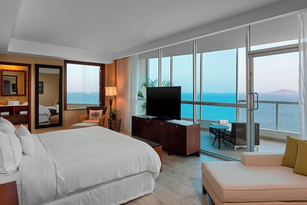 A hotel room with a large bed, a TV, a balcony with a view of the sea, and a seating area with a table and chairs.