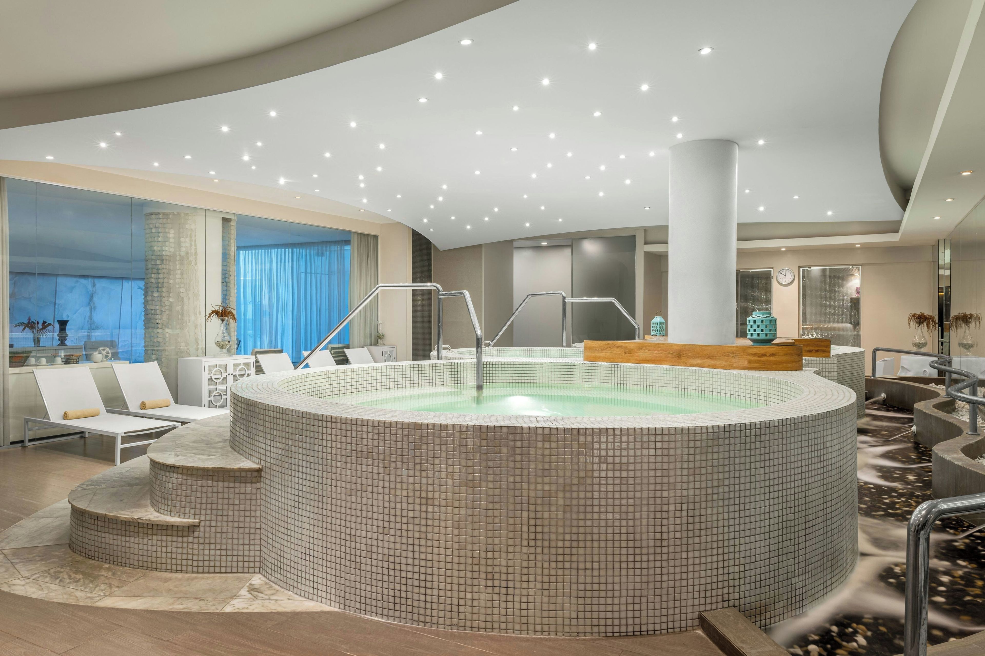 A modern indoor pool area with a hot tub, lounge chairs, and a staircase leading to a higher level.