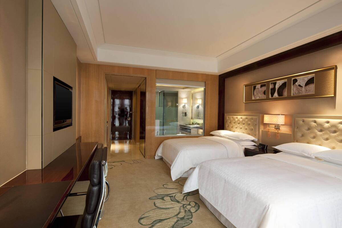 Une chambre d’hôtel avec deux lits, un bureau, une télévision et une salle de bain visible à travers une porte en verre.