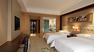 A hotel room with two beds, a desk, a TV, and a bathroom visible through a glass door.