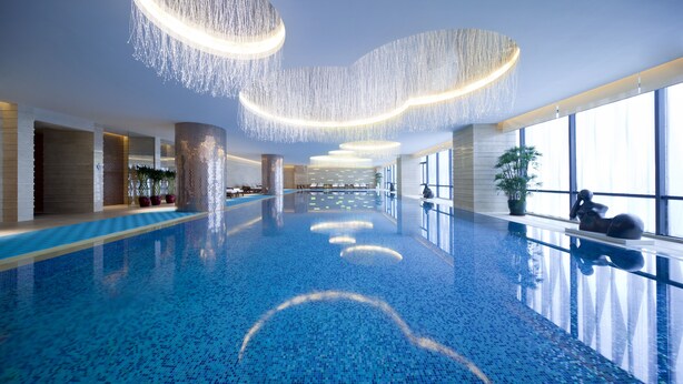 Ein großer, innenliegender Schwimmbereich mit modernem Design, der ein klares, blaues Fliesenmuster im Boden und eine Reihe von runden Leuchten an der Decke zeigt.