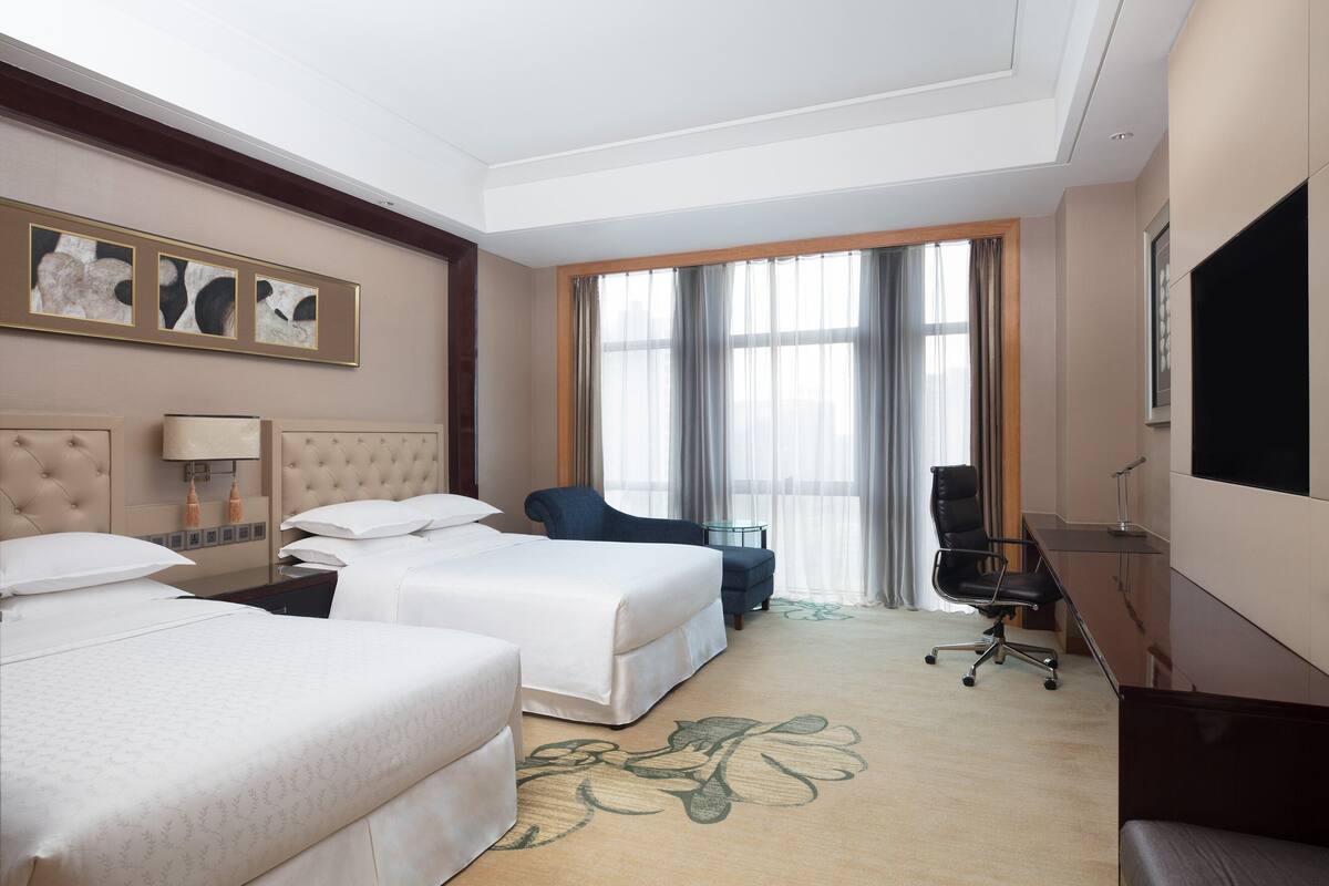 Une chambre d’hôtel avec deux lits, un bureau, une chaise, une télévision et une grande fenêtre avec des rideaux.