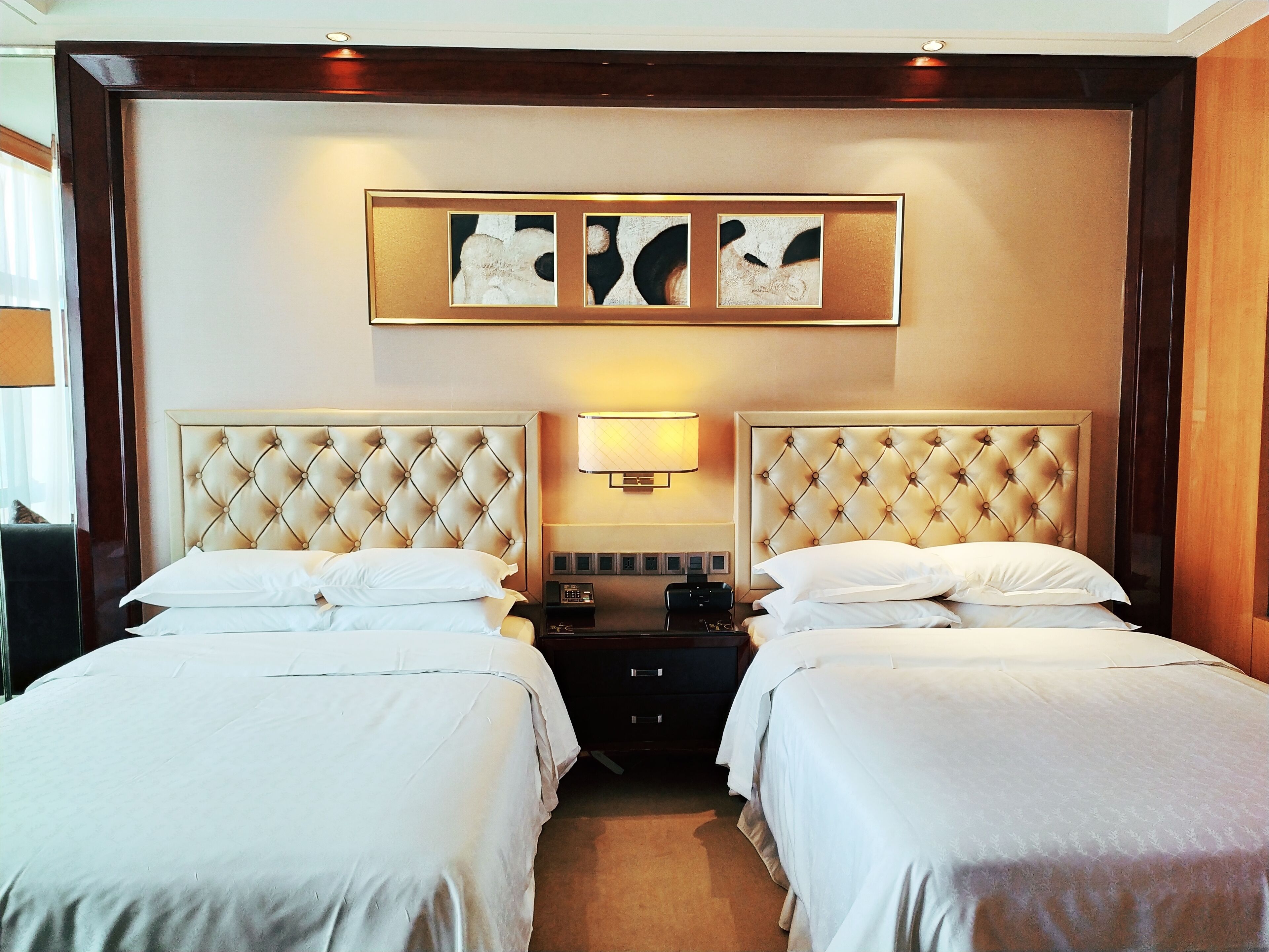 Quarto de hotel com duas camas, uma mesa de cabeceira, uma luminária e uma obra de arte na parede.