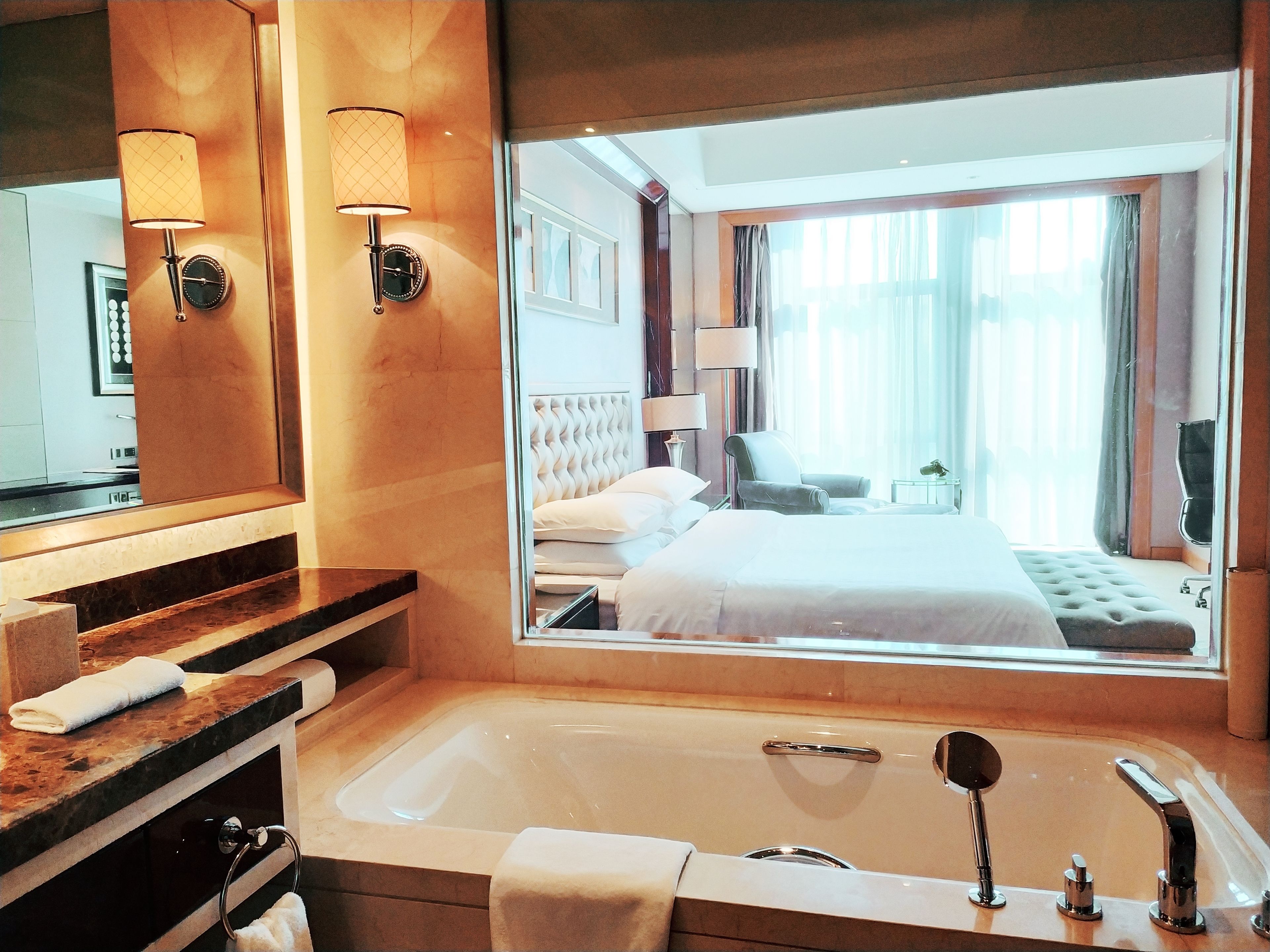 Un baño de hotel con una bañera grande, un espejo y vistas a un dormitorio con cama y sillón.