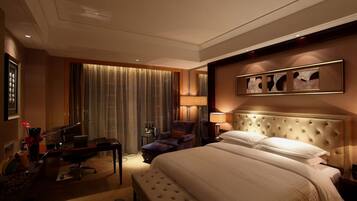 A hotel room with a large bed, a desk with a chair, a small table with a lamp, and a large window with curtains.
