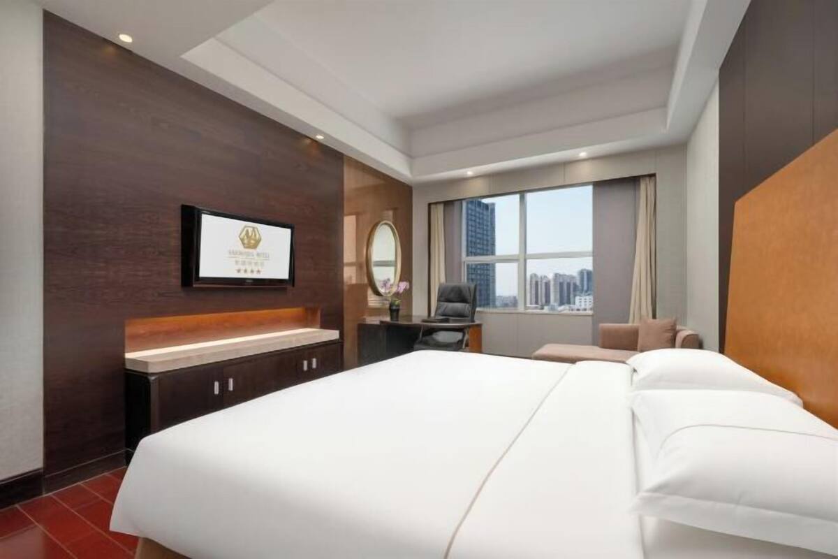 A hotel room with a large bed, a desk, a chair, a TV, and a window with a city view.