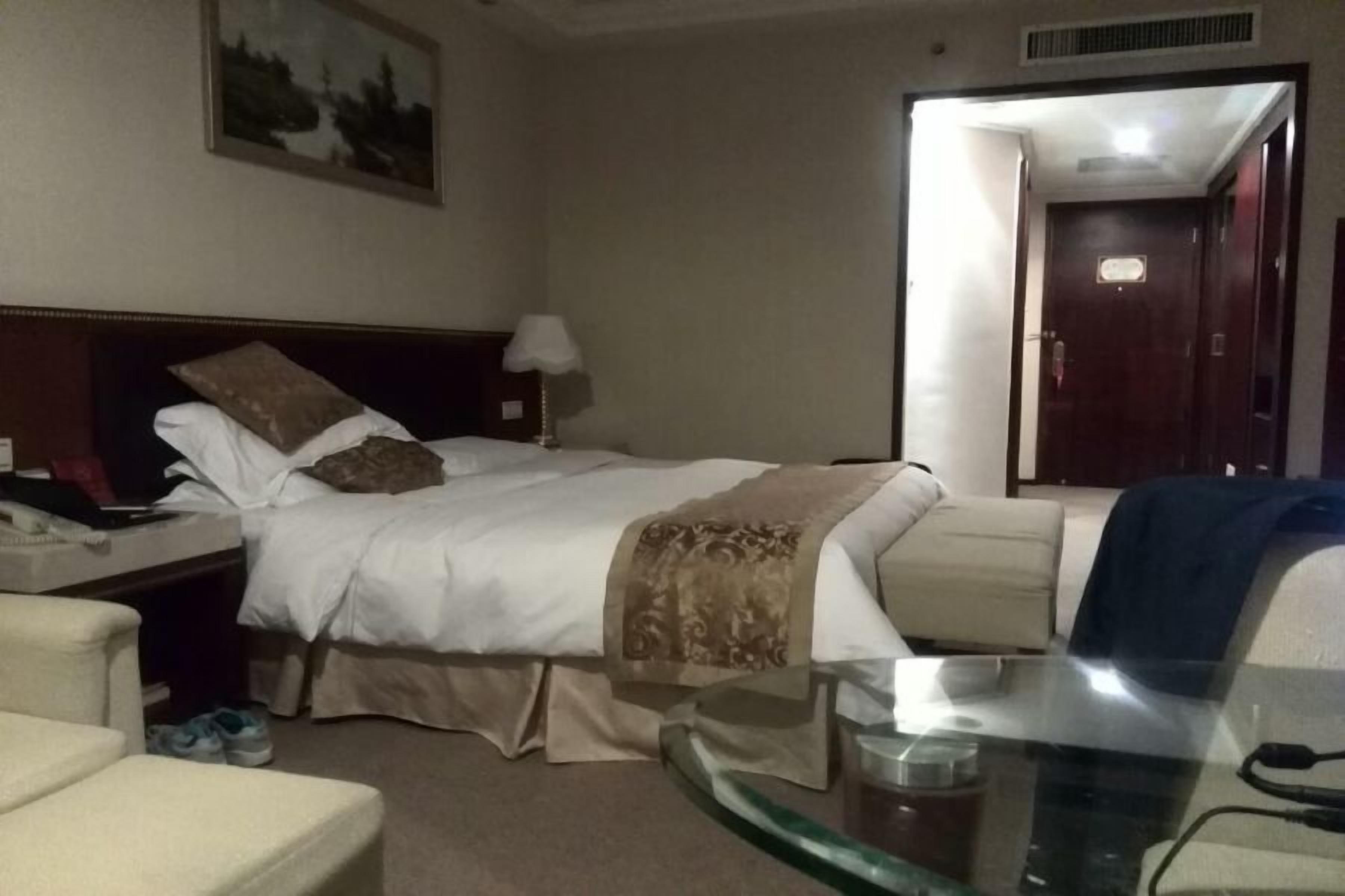 A hotel room with a bed, a bedside table with a lamp, a chair, and a glass table with a cup.