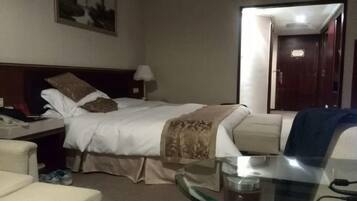 A hotel room with a bed, a bedside table with a lamp, a chair, and a glass table with a cup.