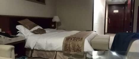Habitación de hotel con una cama, una mesita de noche con lámpara, una silla y una mesa de vidrio con una taza.