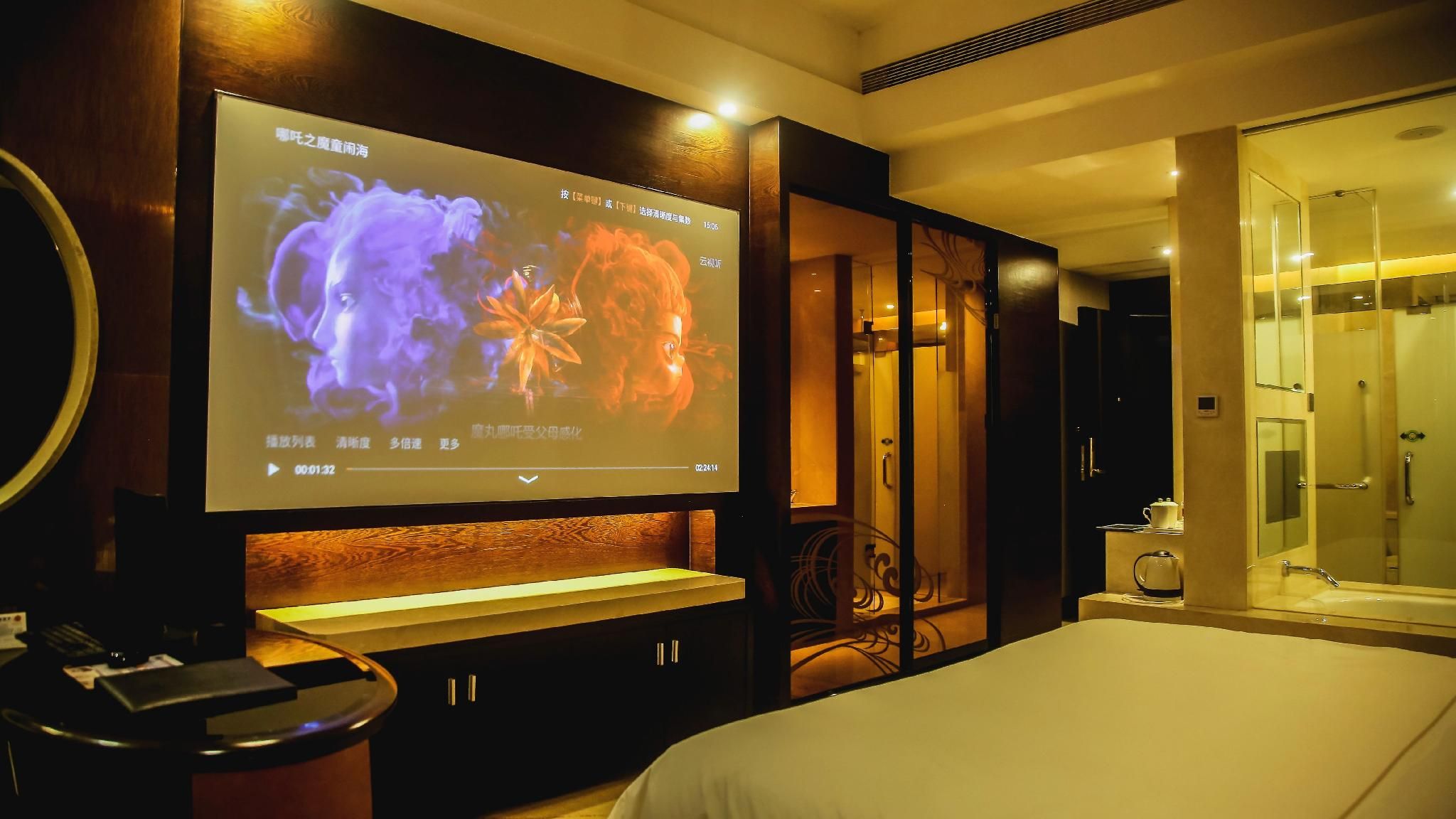 A hotel room with a large flat-screen TV mounted on the wall, a bed, a bedside table, a mirror, and a glass-enclosed shower area.