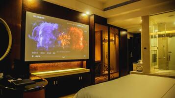 A hotel room with a large flat-screen TV mounted on the wall, a bed, a bedside table, a mirror, and a glass-enclosed shower area.