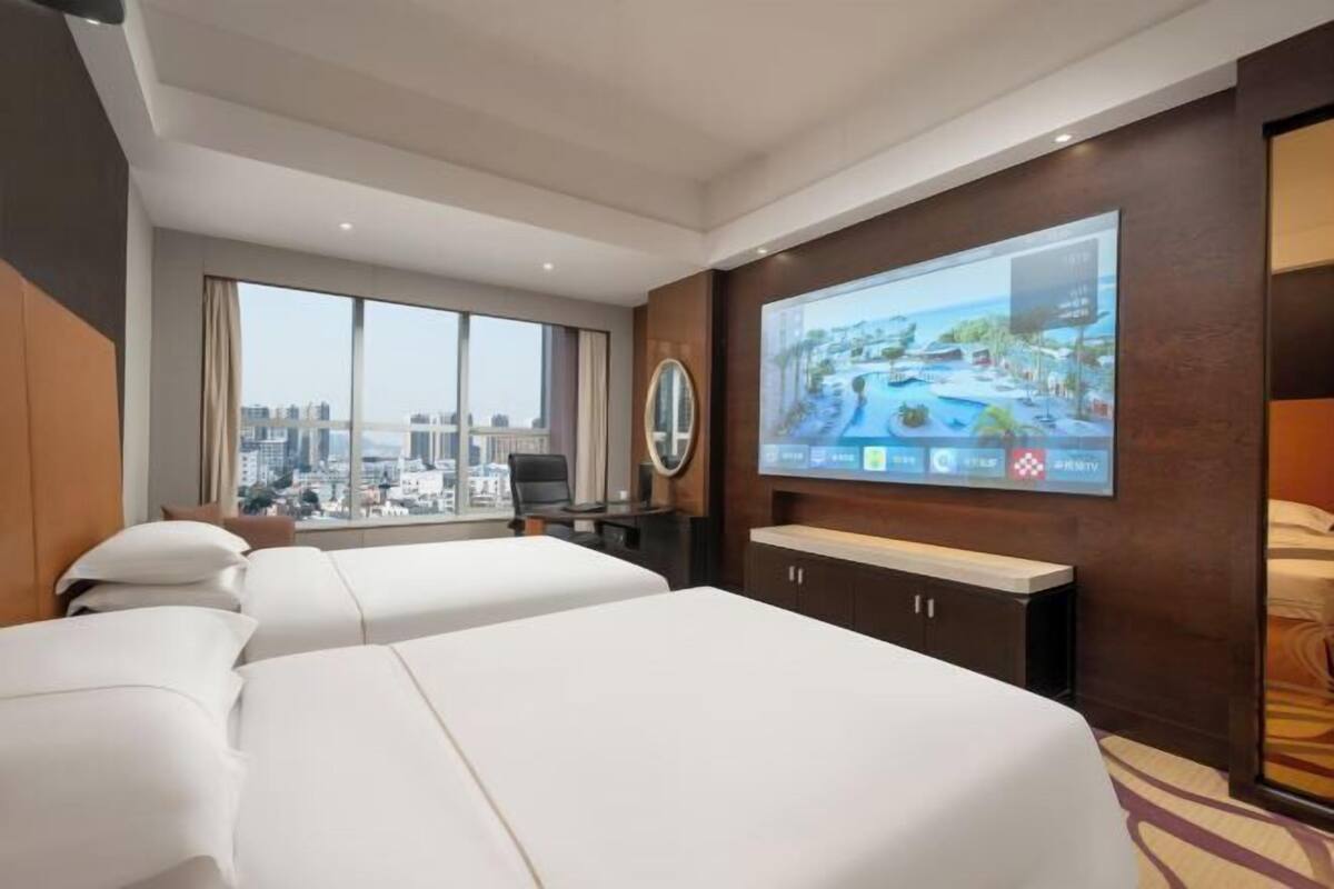 A hotel room with two beds, a large TV screen, a desk, and a view of the city.