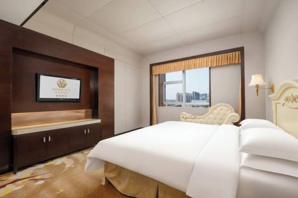 A hotel room with a bed, a mounted television, a window with a city view, and a decorative wall panel.