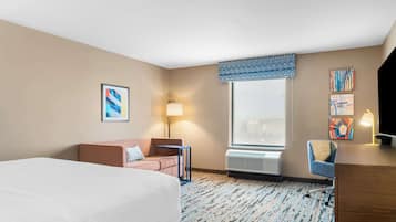 A hotel room with a large bed, a desk, a chair, a sofa, a window with blinds, and a patterned carpet.