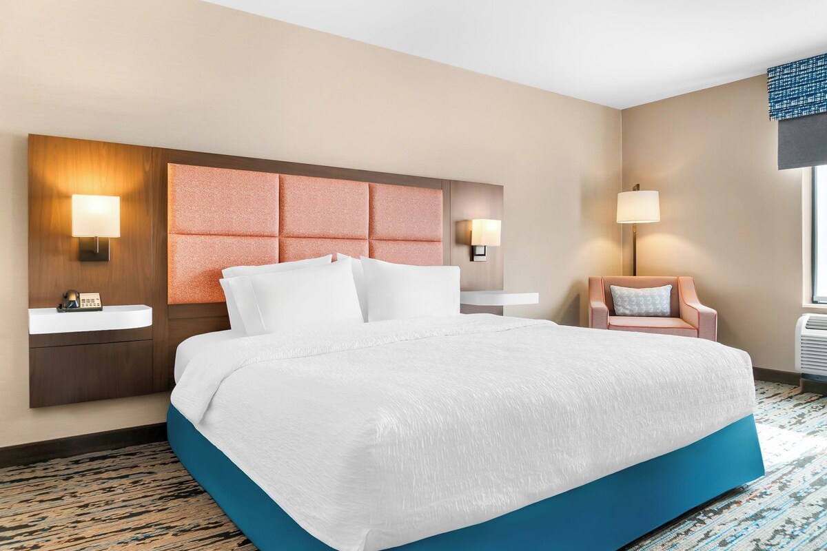 A hotel room with a large bed, a bedside table, a chair, a desk, and a window.