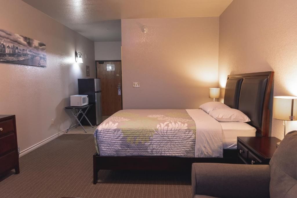 A hotel room with a bed, bedside lamp, a desk, a chair, a TV, and a small table.