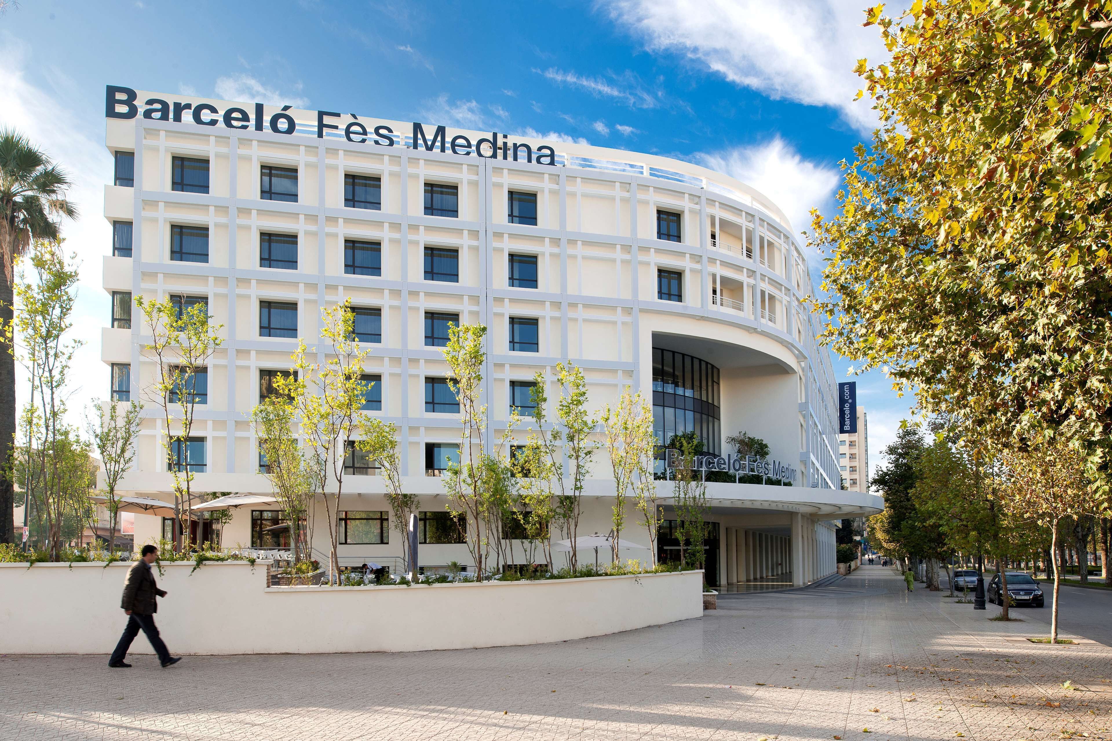 A modern building with a curved facade, named Barceló Fès Medina, featuring a spacious entrance area with trees and outdoor seating.