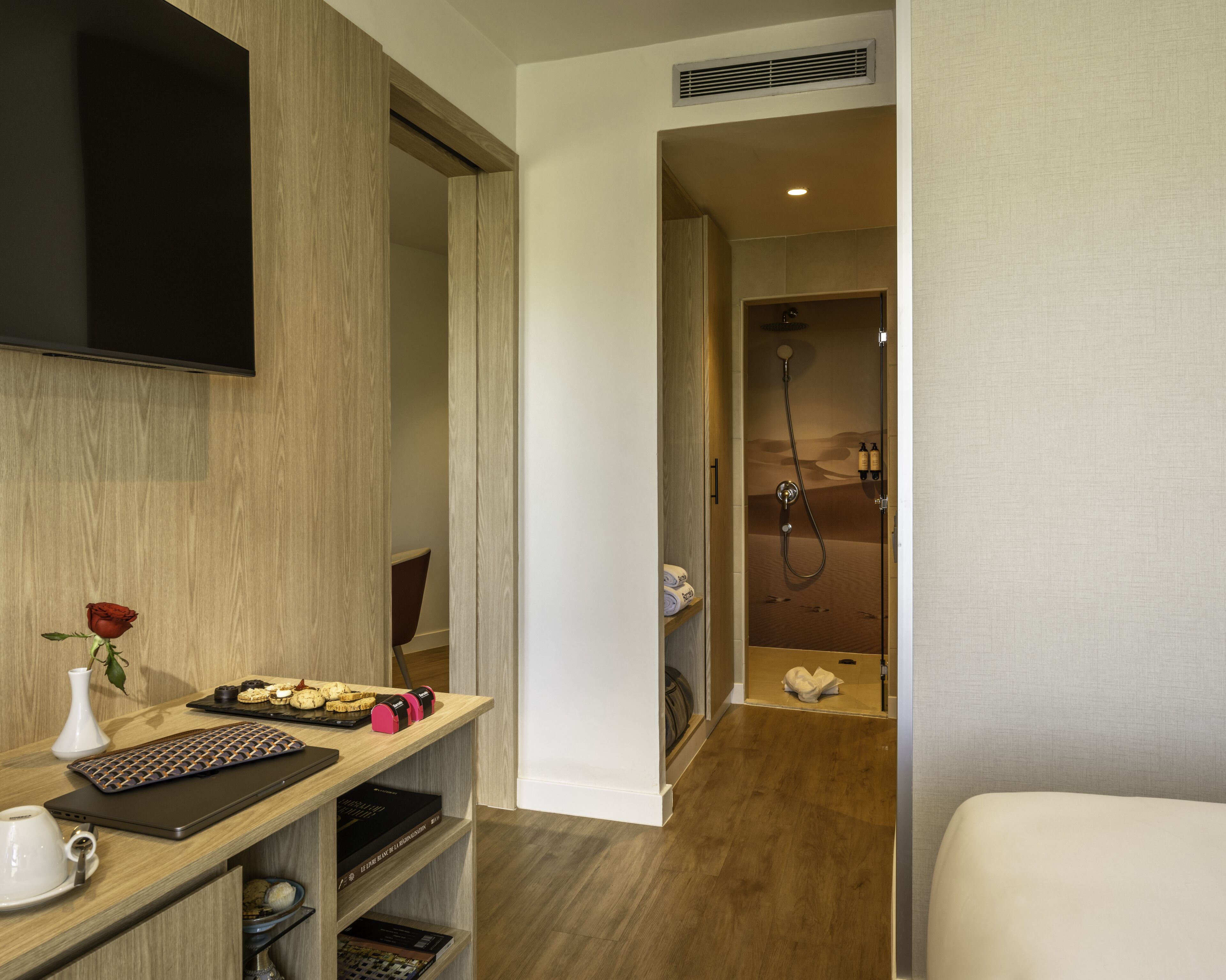 A hotel room with a wooden desk, a laptop, a rose vase, and a shower area with a towel and toiletries.