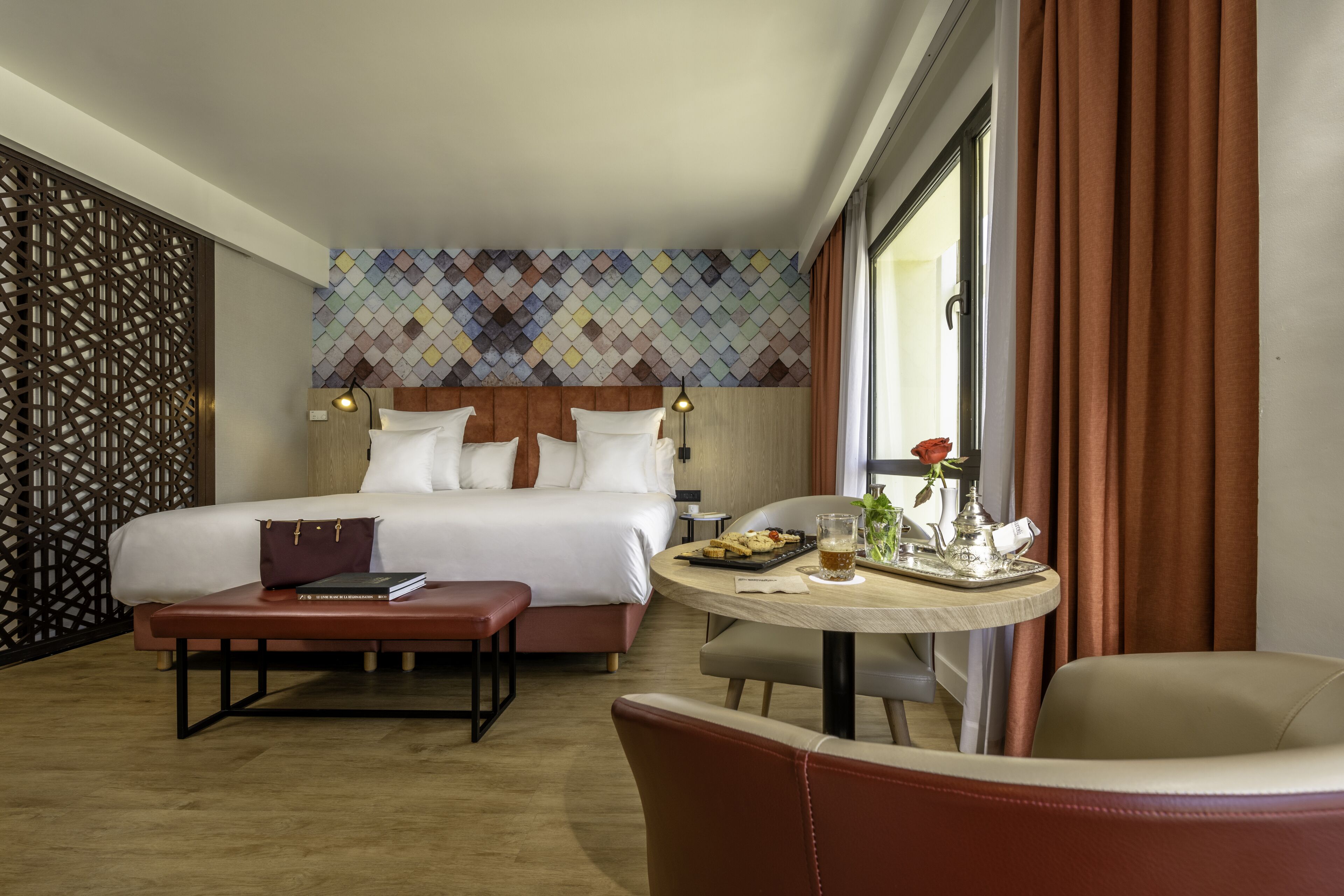 Una habitación de hotel con una cama grande, una mesa redonda con una bandeja de comida, una silla, un banco y un adorno en la pared.