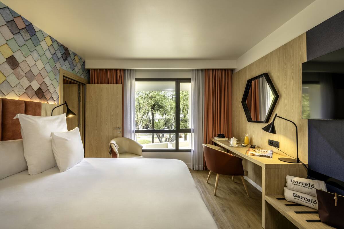A hotel room with a bed, desk, chair, and a view of greenery outside.