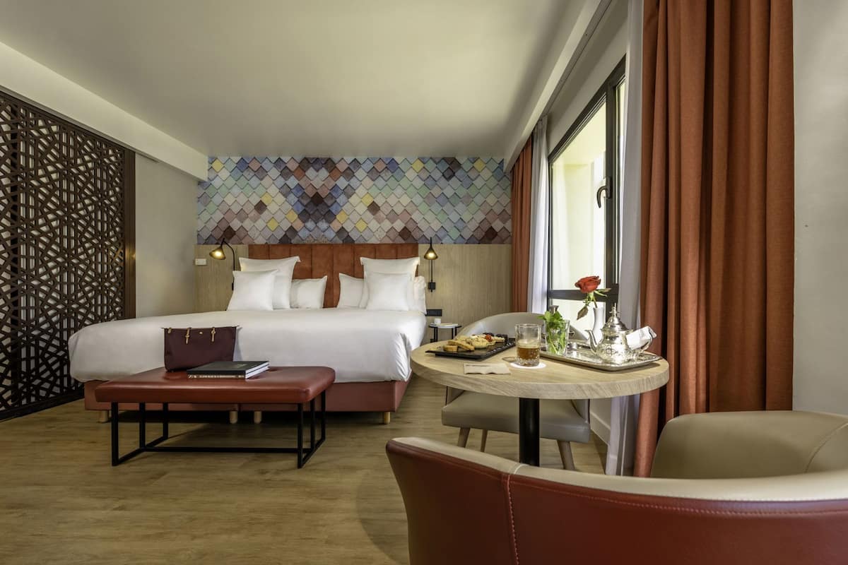 A hotel room with a bed, a dining table with food, a chair, and a window with curtains.