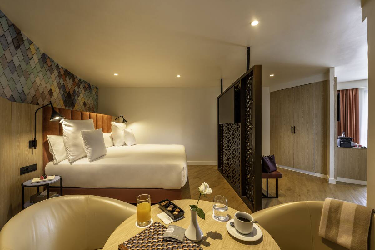 A hotel room with a bed, a table with breakfast items, a coffee cup, and a vase with a flower.