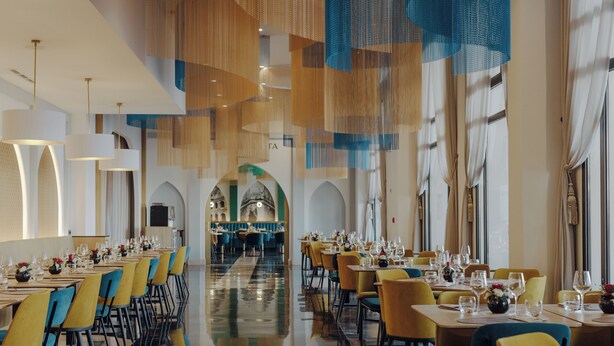 A spacious dining area with multiple tables and chairs, a central walkway, and large windows with curtains. - Barceló Fès Medina (Fes)