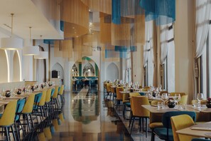 A spacious dining area with multiple tables and chairs, a central walkway, and large windows with curtains. - Barceló Fès Medina (Fes)