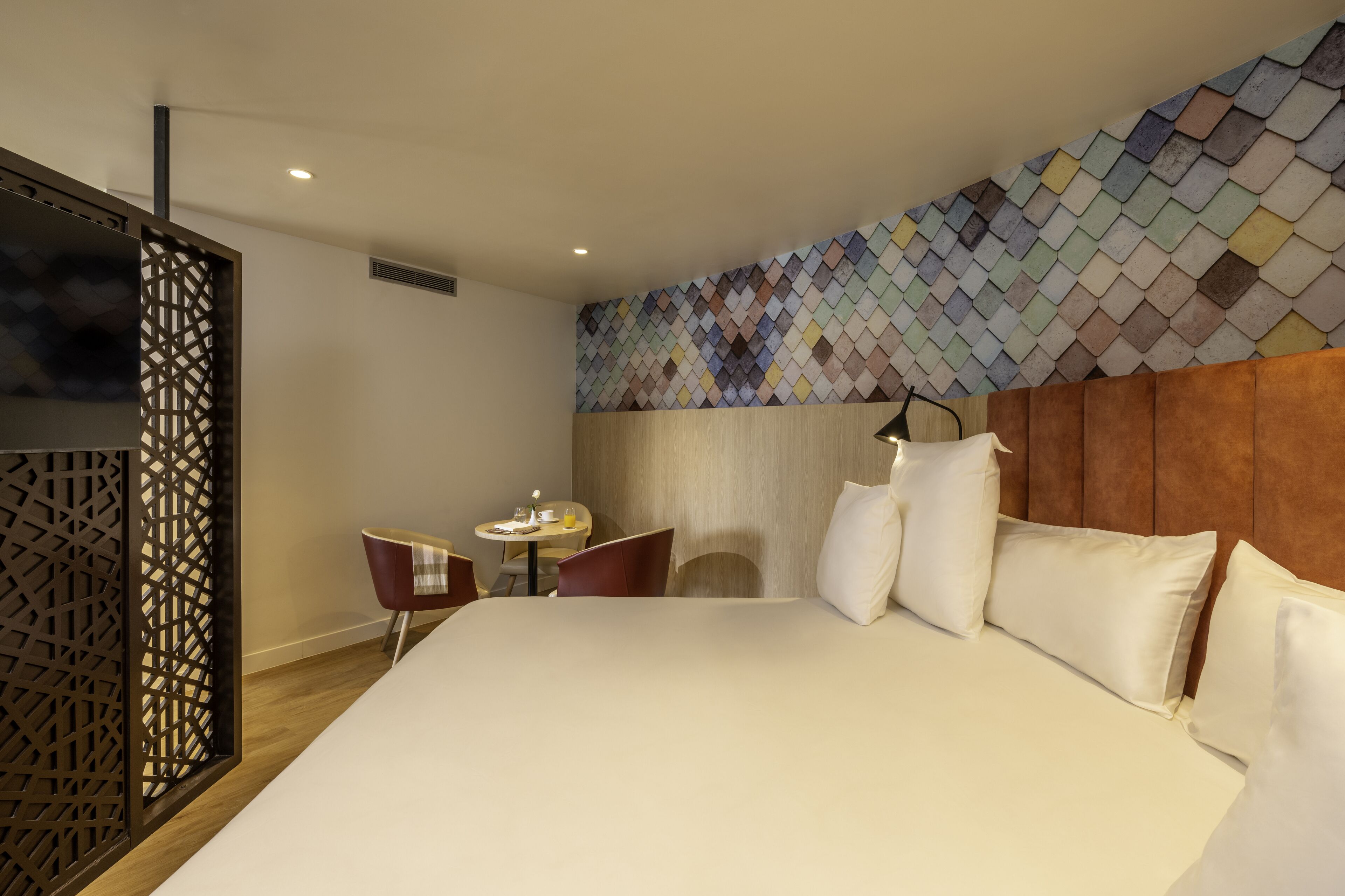 Quarto de hotel moderno com uma cama grande, uma mesinha redonda com duas cadeiras e uma parede decorativa com padrão geométrico.