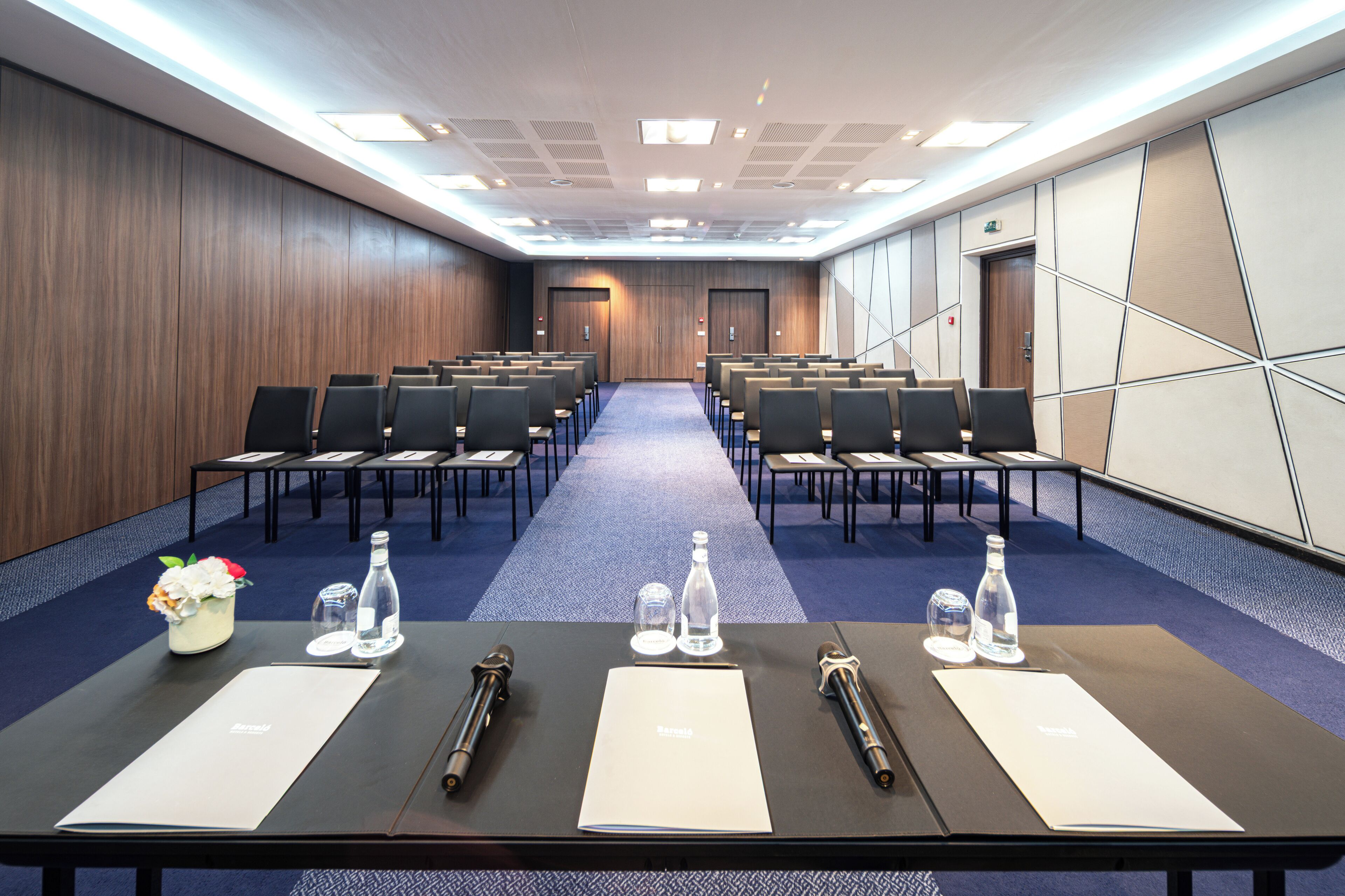 A conference room with rows of chairs, a long table with nameplates and water bottles, and a wooden wall.