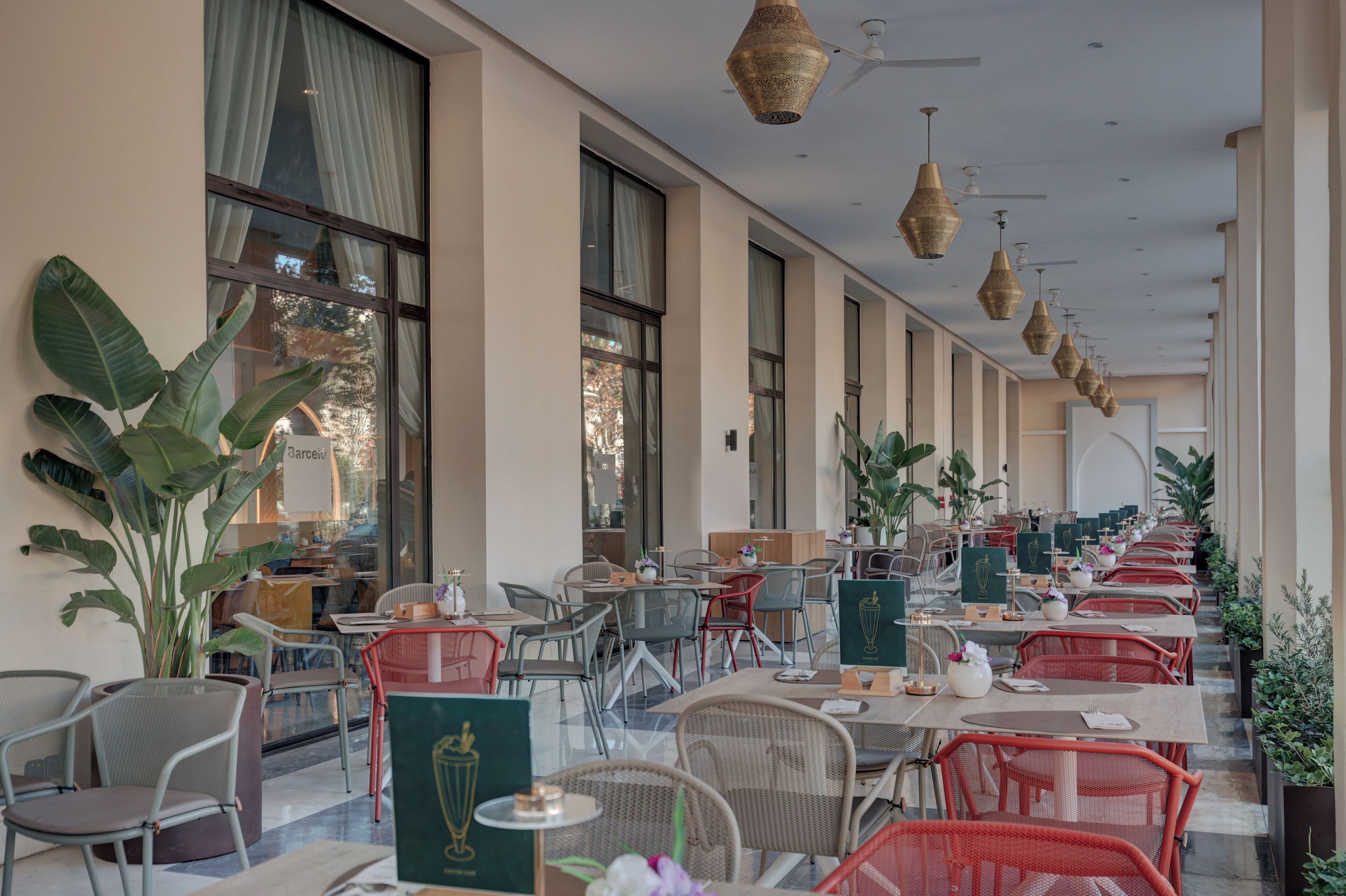 A long, covered outdoor dining area with tables and chairs, large windows, and hanging light fixtures.