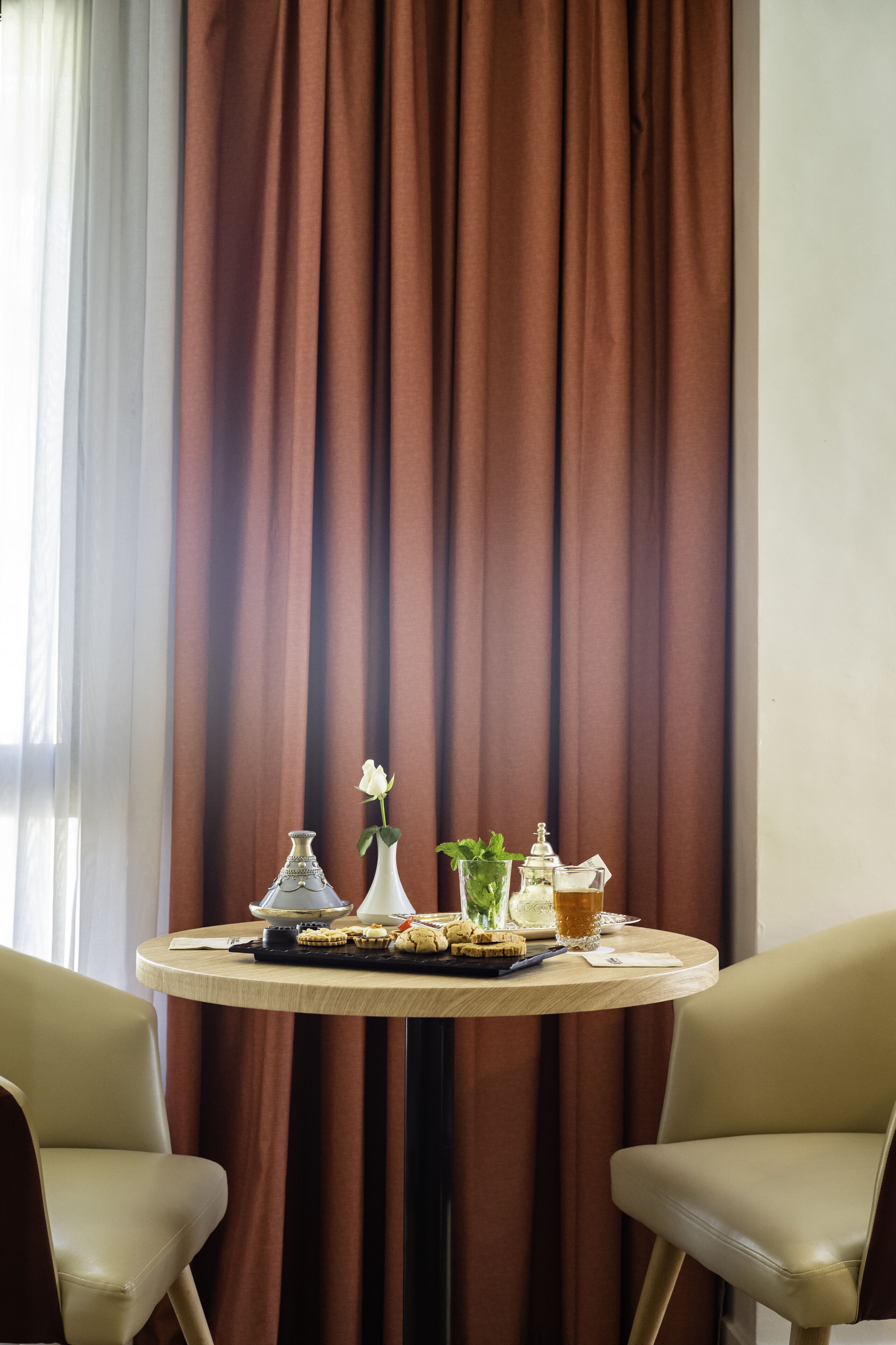 Una mesa redonda con un florero blanco, una rosa blanca, un recipiente de vidrio y una planta pequeña. Dos sillas color beige están ubicadas a cada lado de la mesa.