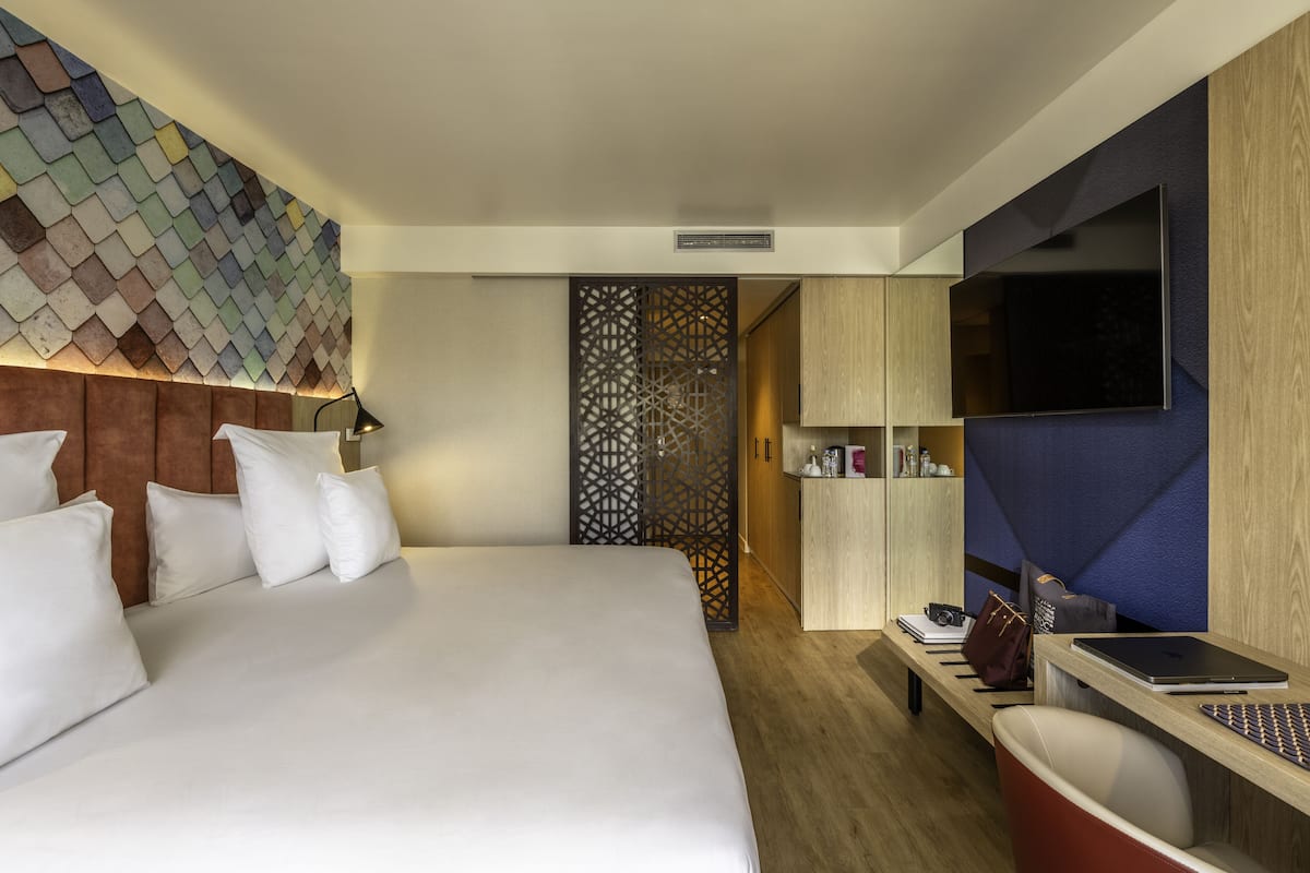 A hotel room with a large bed, a TV mounted on the wall, a desk with a laptop, and a decorative wall panel.