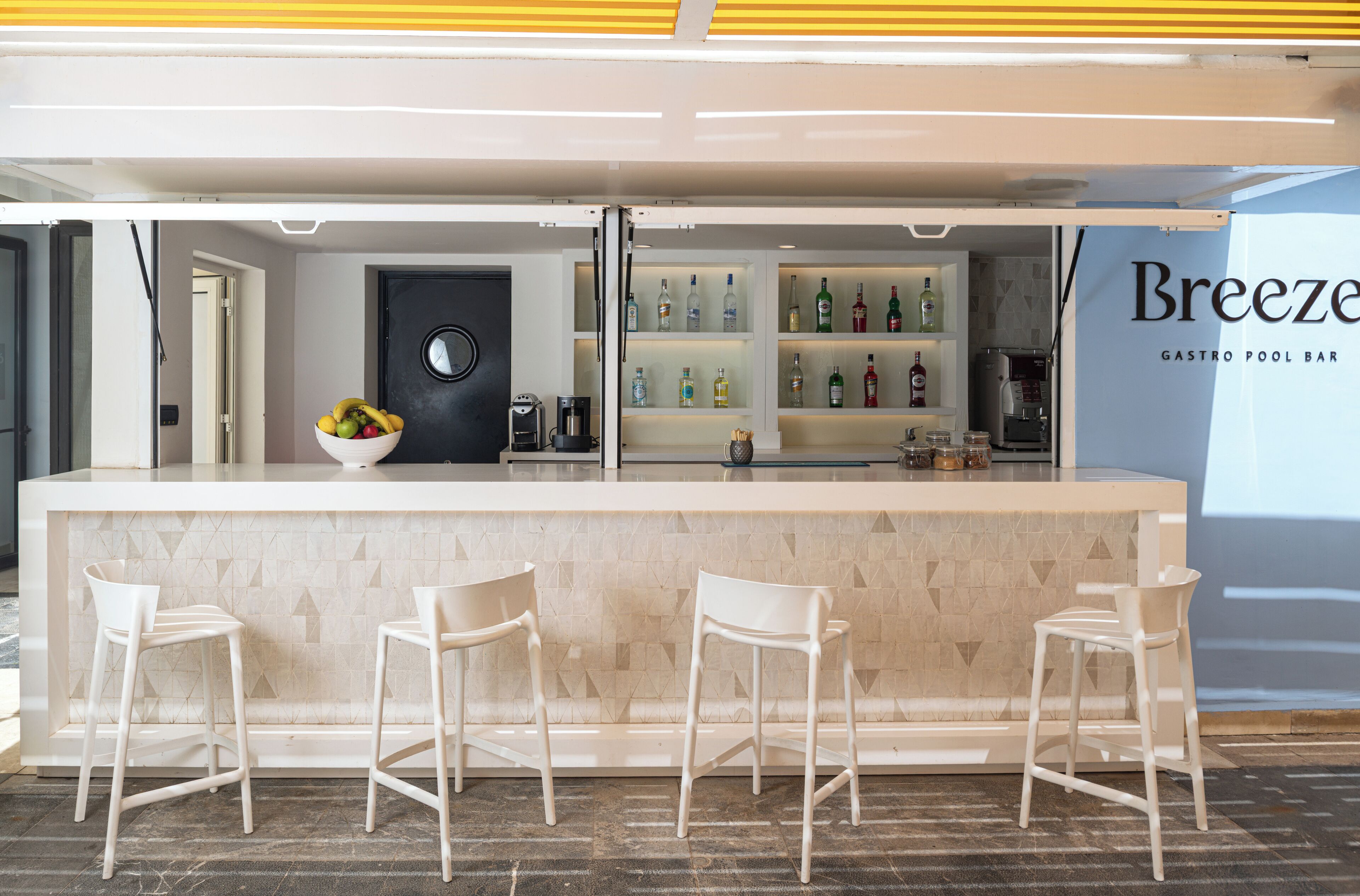 Zona de bar con mostrador de mármol, taburetes blancos, un cuenco de fruta y estantes llenos de botellas.