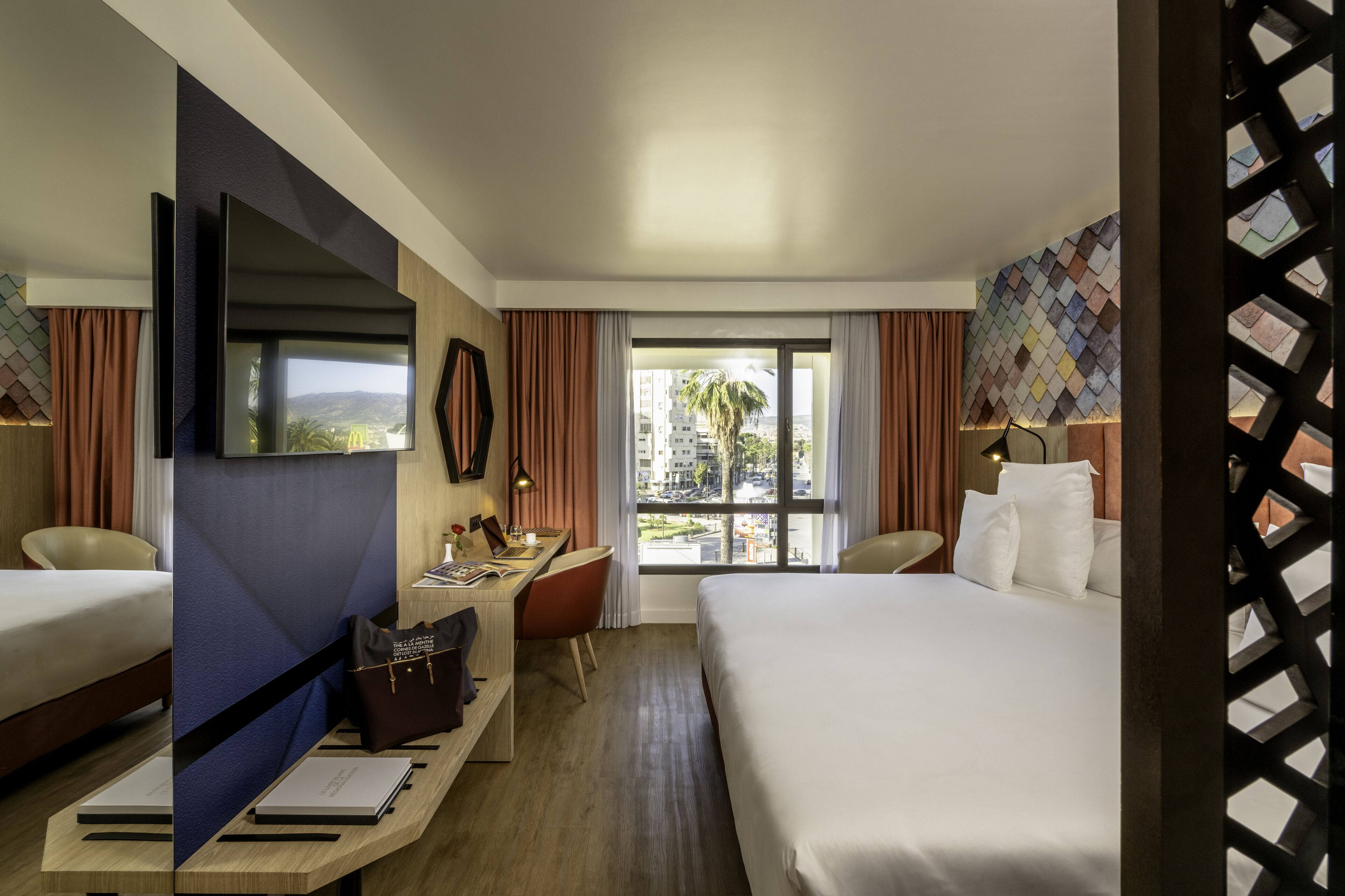 A hotel room with two beds, a desk, a TV, and a view of the city.