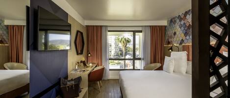 A hotel room with two beds, a desk, a TV, and a view of the city.
