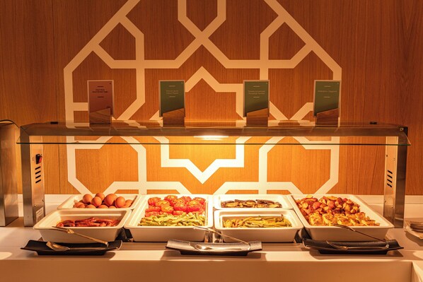 A buffet with various food options, including fruits, vegetables, and prepared dishes, displayed in white trays with labels.
