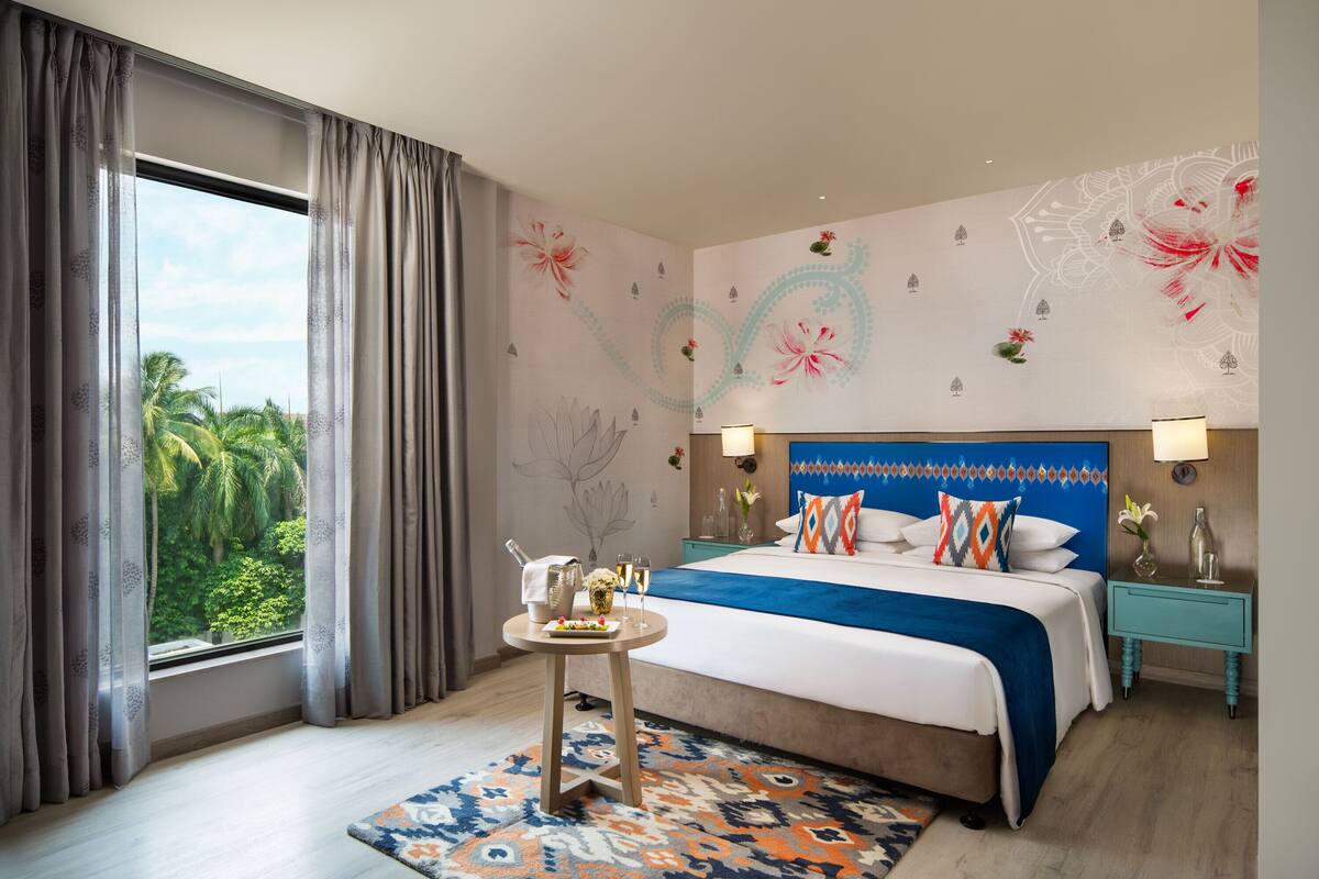 A hotel room with a large bed, a bedside table, a nightstand, and a view of palm trees through the window.