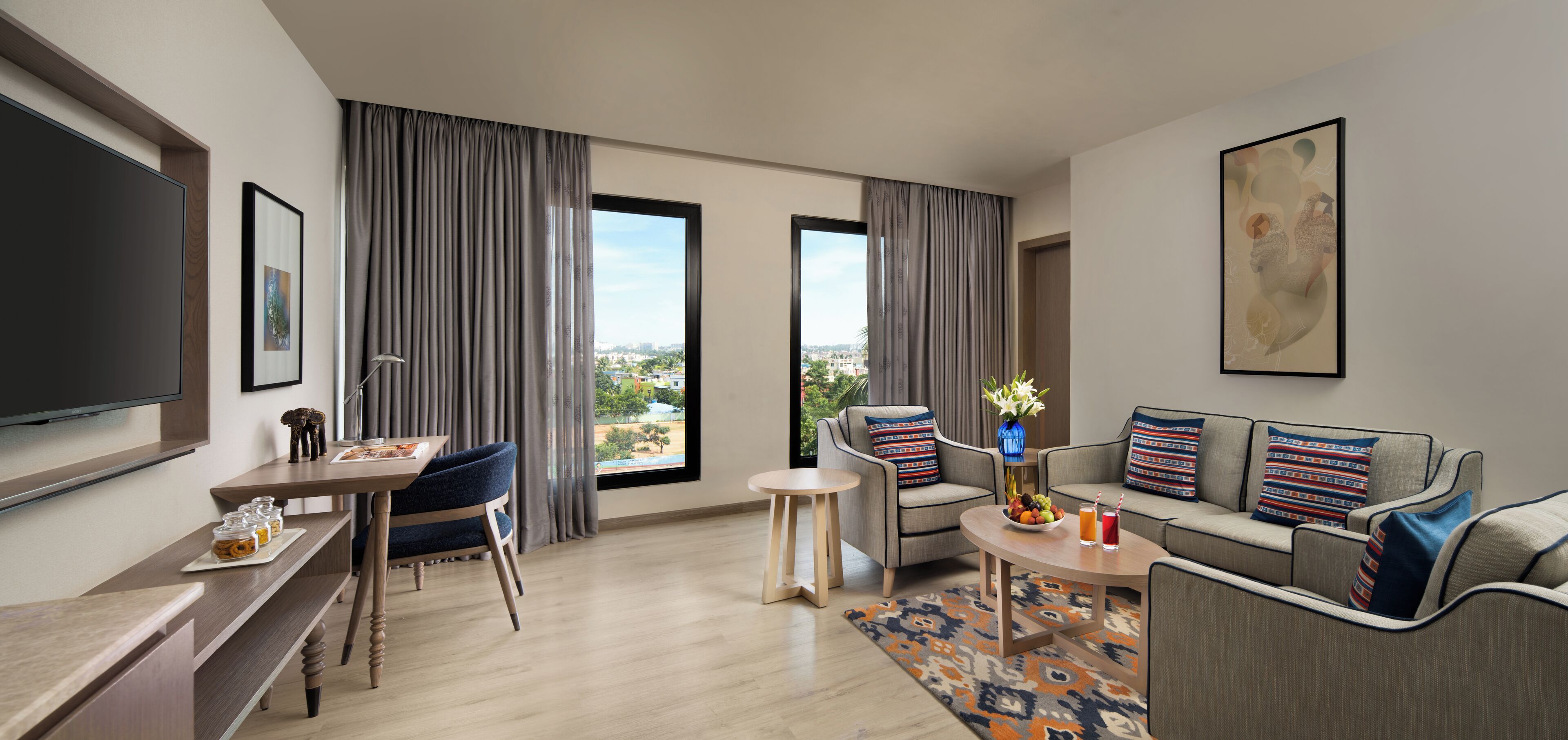 Une chambre d’hôtel moderne dotée d’une grande télévision à écran plat, d’un ensemble de canapés, d’un coin repas avec une table et des chaises, et d’une grande fenêtre offrant une vue sur la ville.