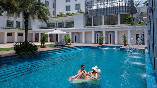 Une piscine où un couple se détend sur un flotteur, entouré de palmiers et d’un bâtiment moderne doté de balcons.