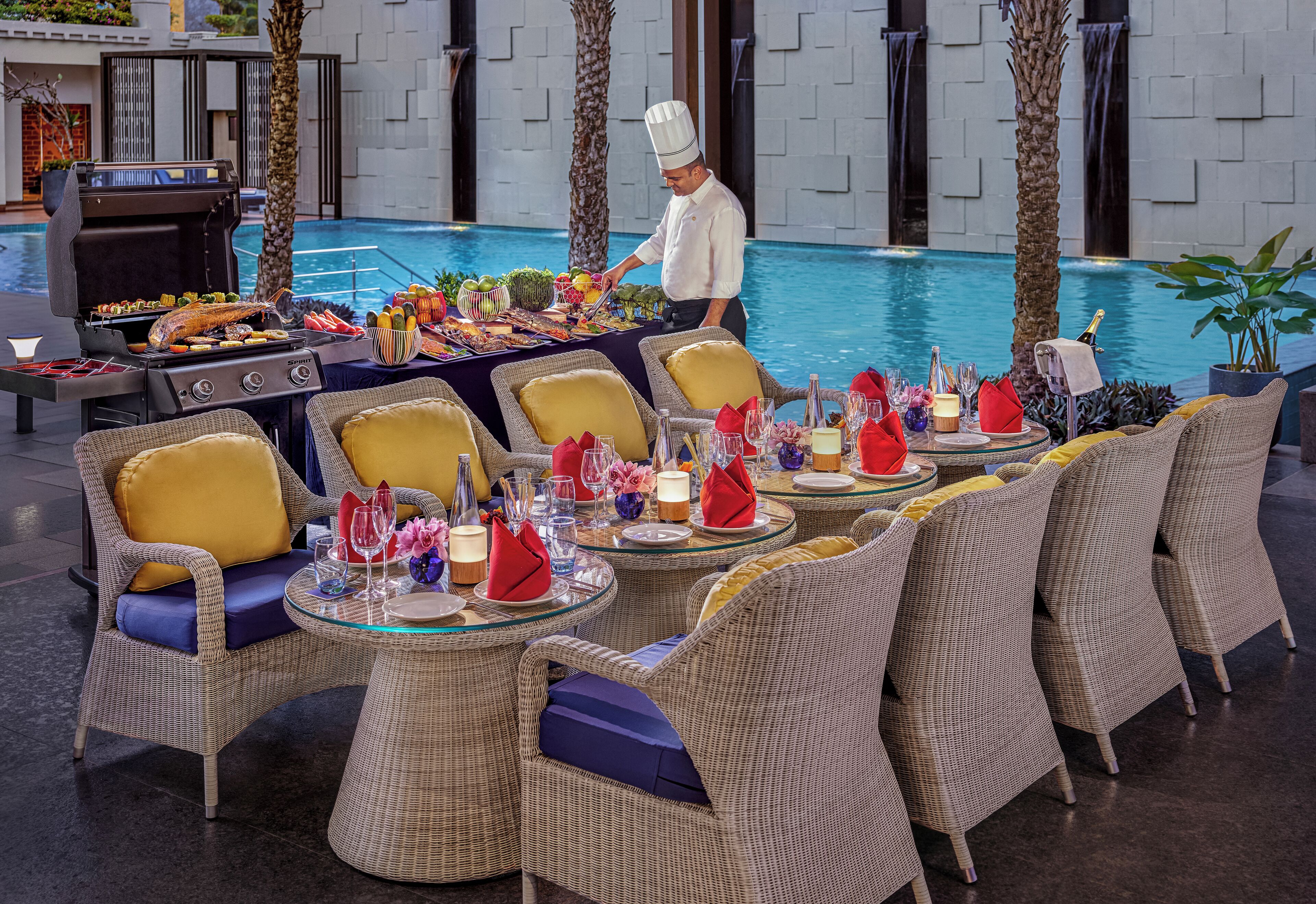 An outdoor dining area with wicker chairs and tables, a grill with food, and a pool in the background.