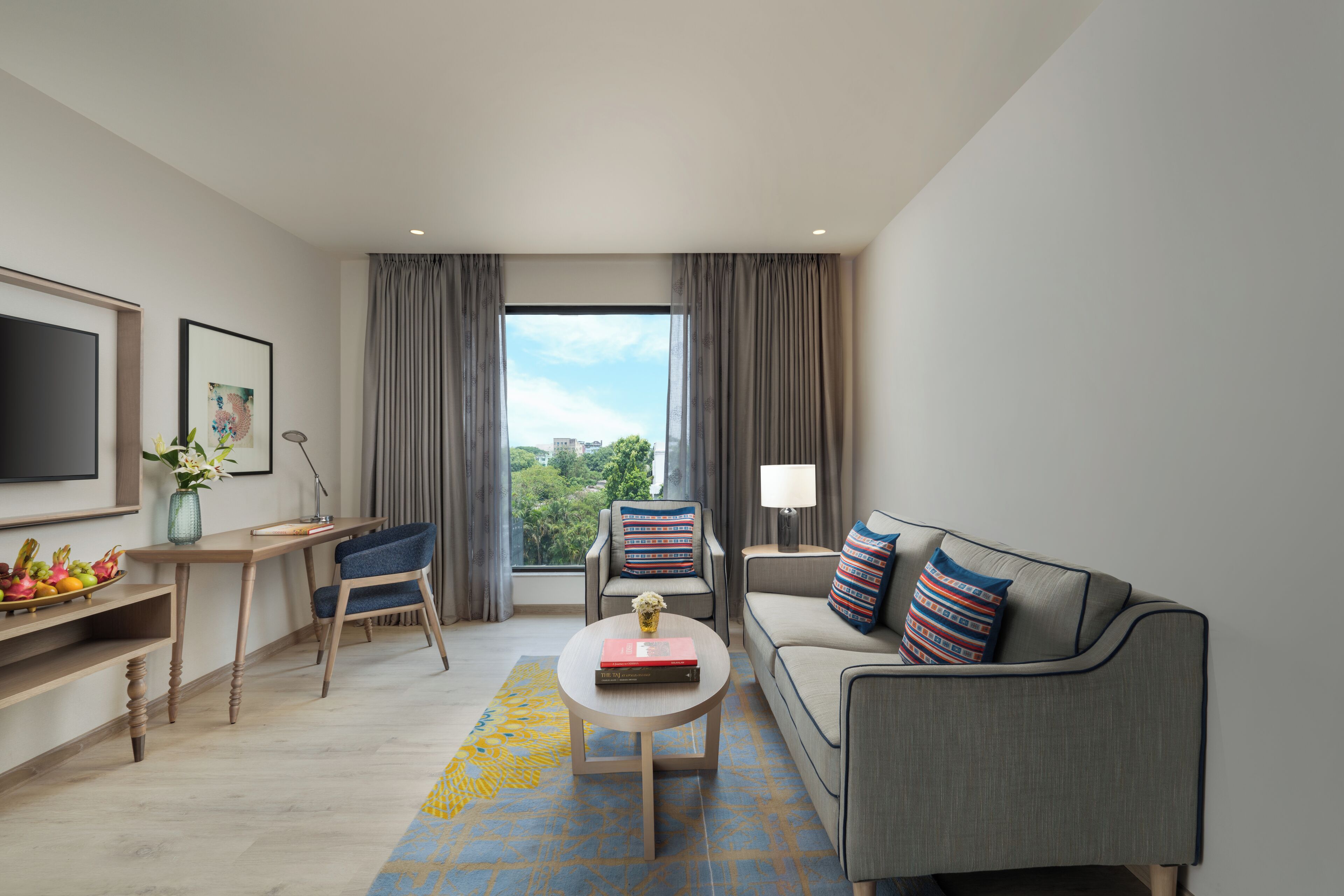 A modern hotel room with a sofa, a chair, a coffee table, a TV, and a large window with curtains.