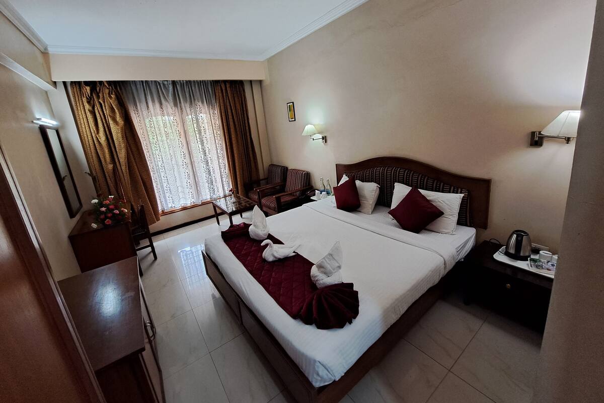 A hotel room with a large bed, a desk, a chair, a nightstand, a lamp, and a window with curtains.