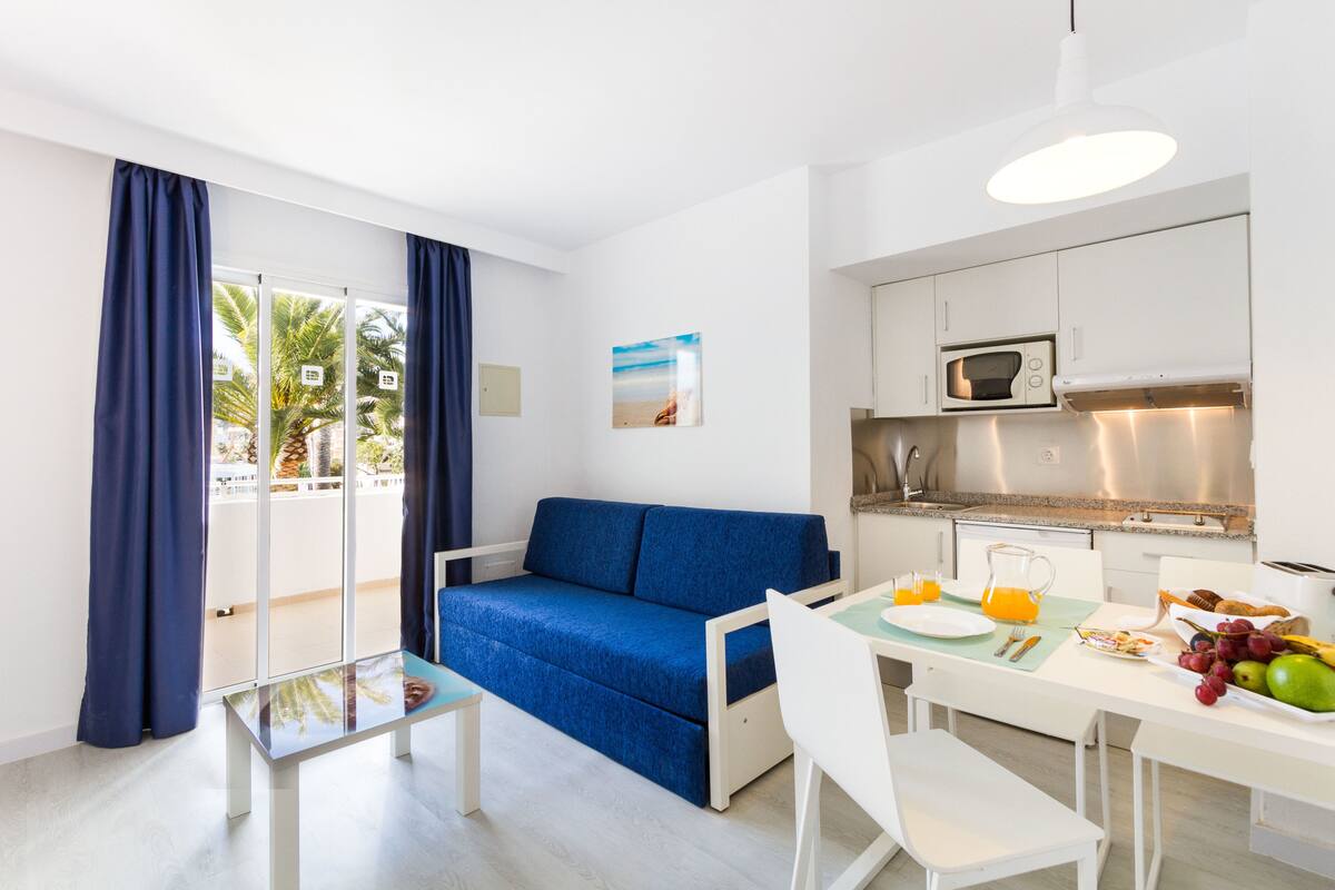 A compact living space with a sofa, a dining table with breakfast items, a kitchen area, and a view of palm trees through the window.