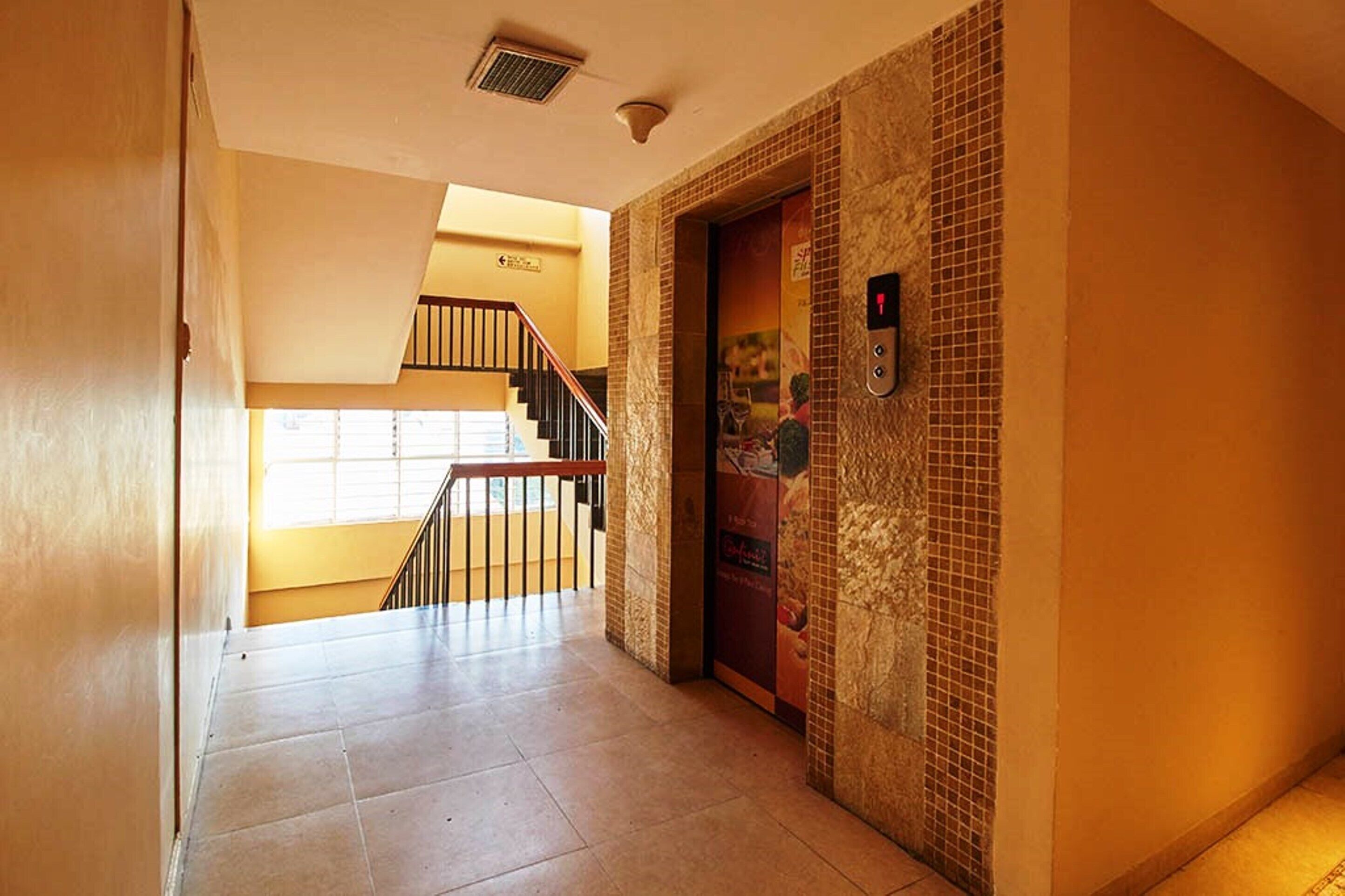 A hotel hallway with a tiled floor, a staircase with a metal railing, and a wall-mounted elevator control panel.